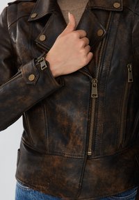 Giacca da moto in pelle marrone vissuta con accessori in argento, tasche con zip e dettaglio a bottone sui polsini. Design testurizzato e aderente.