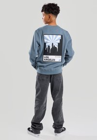 Sudadera azul con un gráfico de paisaje urbano en negro y azul claro, que incluye el texto "LOS ÁNGELES". Emparejada con jeans grises y zapatos negros.