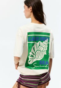 Cream-colored oversized t-shirt with a green graphic featuring a seashell and text. Short sleeves, relaxed fit, soft cotton material.