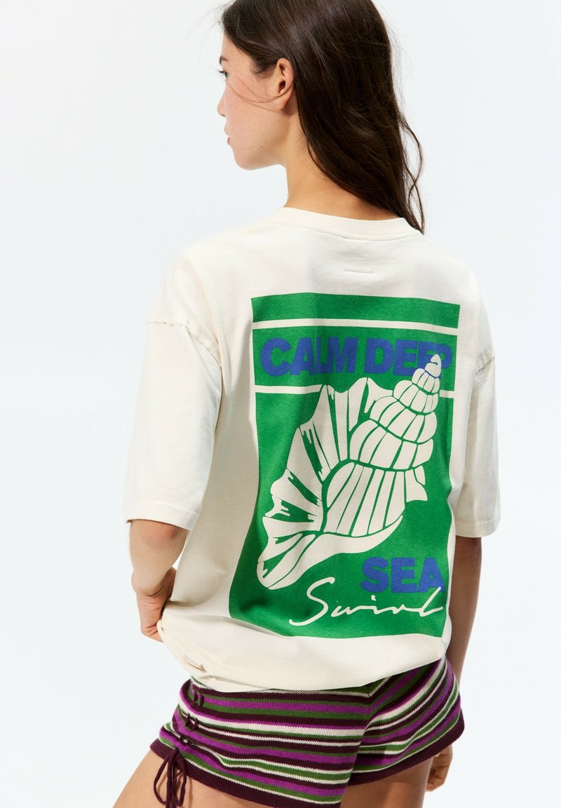 Cream-colored oversized t-shirt with a green graphic featuring a seashell and text. Short sleeves, relaxed fit, soft cotton material.