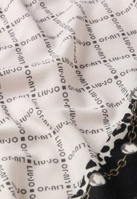 Tissu blanc avec le texte noir "LIU·JO" répété en motif quadrillé, froissé, montrant un détail de design en chaîne et perles en noir et blanc.