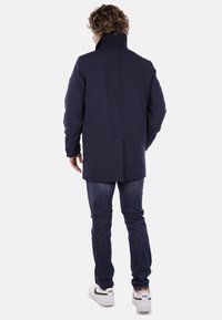 Giacca da esterno blu navy con colletto alto, maniche lunghe e taglio dritto. Indossata con jeans scuri e sneakers bianche. Tessuto liscio.