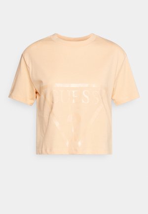 Lichtperzikkleurig cropped T-shirt met korte mouwen en een subtiel glanzend Guess-logo en vraagteken aan de voorkant tegen een effen achtergrond.