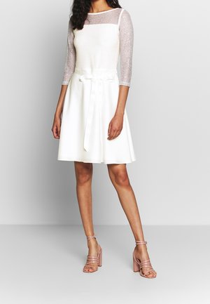 Biała sukienka z dopasowanym gorsetem i rozkloszowanym dołem, ozdobiona przezroczystymi, błyszczącymi długimi rękawami oraz atłasowym paskiem w talii. Różowe sandały na obcasie z paskami.