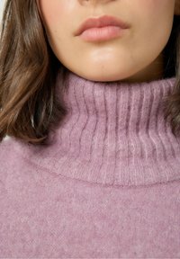 Šviesiai rožinė megztinis su aukštu raštuotu apykakle, pasižymintis minkšta tekstūra ir subtiliu mezgimo rašto skirtumu. Artimas vaizdas, sutelkiantis dėmesį į apykaklę.