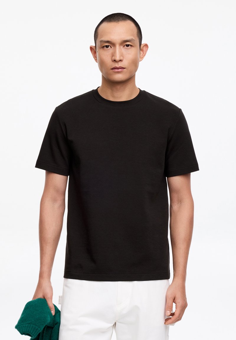 Homme aux cheveux courts portant un t-shirt noir et un pantalon blanc, tenant un pull vert plié dans sa main gauche sur fond blanc.