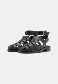 Sandali in pelle nera con più cinghie orizzontali, fibbia alla caviglia, suola piatta e design aperto sulla punta. Texture liscia con accentuazioni minimal.
