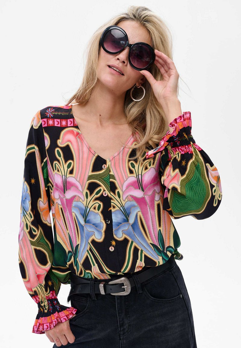 Kobieta w dużych czarnych okularach przeciwsłonecznych, kolorowej kwiecistej bluzce z bufiastymi rękawami, z okrągłymi kolczykami, czarnym paskiem i ciemnymi dżinsami, pozująca na tle białym.