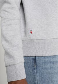 Felpa grigia chiara con polsini, ornata da piccoli dettagli ricamati rossi, bianchi e blu, abbinata a jeans in denim azzurro.
