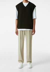 Černý svetřík bez rukávů s výstřihem do V nošený přes bílé tričko, kombinovaný s béžovými širokými kalhotami a bílými teniskami. Hladká látka, ležérní design.