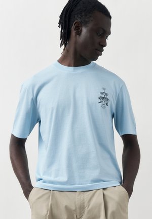 Άνδρας με σκούρο δέρμα και dreadlocks που φορά φωτεινό μπλε μπλουζάκι με μικρό μαύρο σχέδιο κοκτέιλ στο στήθος και μπεζ παντελόνι.