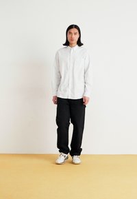White button-up shirt with vertical stripes, black loose-fit trousers, standing on a light-colored floor. Basic white sneakers complete the look.