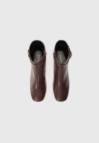 Bottines en cuir marron avec une texture lisse, bout rond et fermeture à glissière sur le côté. Accent en matériel doré sur les côtés.