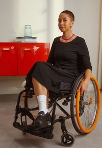 Femme en robe noire assise dans un fauteuil roulant aux roues orange, à l'intérieur près d'un cabinet rouge avec des livres et un bocal en verre sur le dessus.