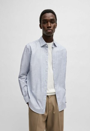 Chemise à boutons en bleu clair et blanc à rayures verticales, fabriquée en coton. Équipée d'un col classique et de manches longues, associée à un pantalon beige.