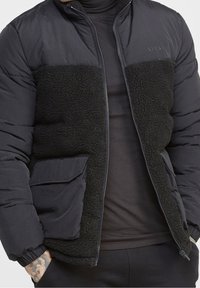 Svart puffjacka med en fluffig svart fleeceframpanel, dragkedja och elastiska ärmslut. Har en verktygsficka på vänster sida.