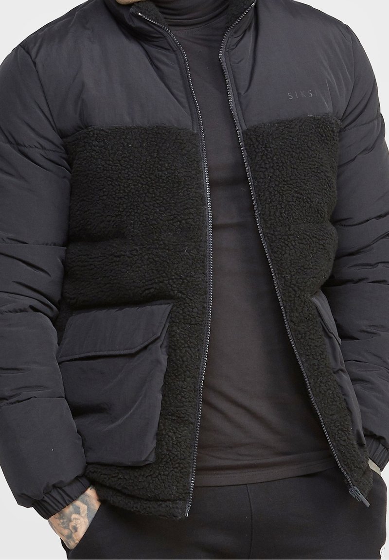 Svart puffjacka med en fluffig svart fleeceframpanel, dragkedja och elastiska ärmslut. Har en verktygsficka på vänster sida.