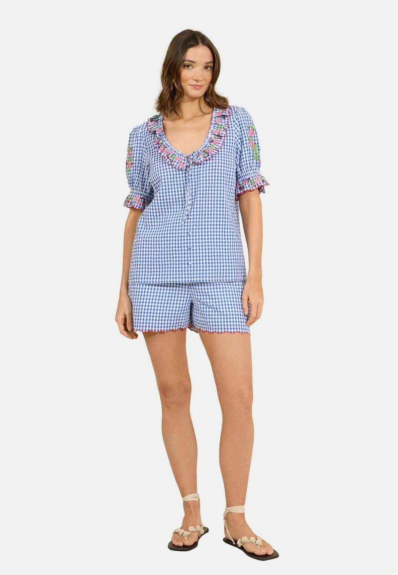 Femme portant un haut à manches courtes bleu et blanc à carreaux avec des volants floraux et un short assorti, debout en sandales beiges sur fond blanc.