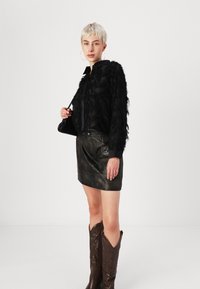Giacca nera con frange, materiale trasparente, abbinata a una minigonna di pelle nera. Stivaletti cowboy marroni con cuciture dettagliate. Borsa semplice.