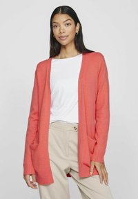 Korallefarbene Strickjacke mit offenem Vorderdesign, gerippten Bündchen und zwei seitlichen Taschen, getragen über einem weißen Hemd und beigen Hosen.