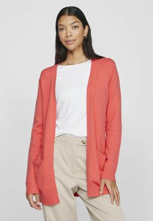 Koraal geklede cardigan met een open voorkant, geribbelde manchetten en twee zijzakken, gedragen over een witte blouse en beige broek.