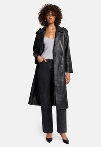 Cappotto trench in pelle nera con design doppiopetto, ampi rever e due tasche frontali. Indossato sopra un top in maglia grigia e jeans scuri.
