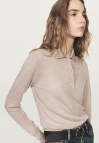 Jasnoróżowa koszulka polo z długim rękawem, kołnierzykiem oraz czterema guzikami, o miękkiej fakturze. Połączona z ciemnym paskiem i dżinsami.