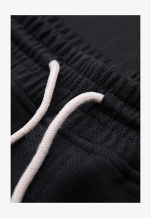 Primer plano de la cintura de unos pantalones deportivos negros con cordón blanco atravesado por una banda elástica acanalada.