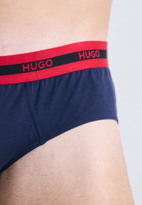 Morske bokserice s rdečo in črno elastično vbočeno pasom, ki vsebuje besedilo "HUGO". Mehka tkanina z gladko teksturo in prileganjem.