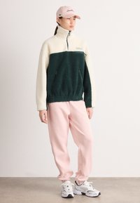 Flis pulover s blokovima boja zelene i krem, polu-zip, bočne džepove; uparen s ružičastim trenerkama i bijelim sportskim cipelama.