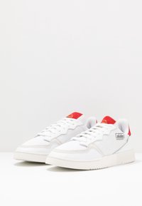Pair of white Adidas Supercourt sneakers with red heel tabs and tongue accents, placed on a white surface with a plain background.