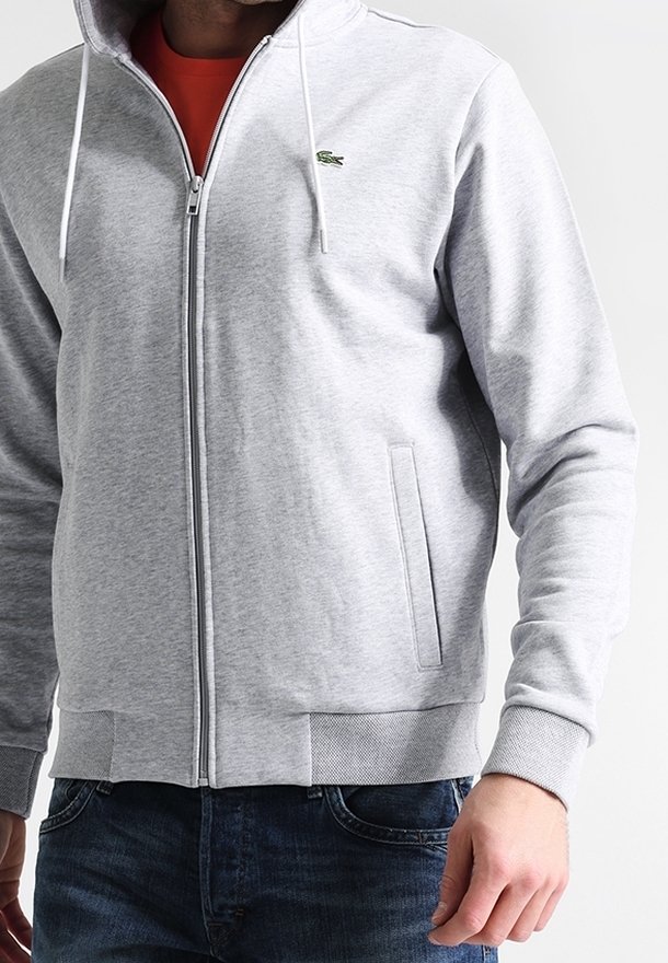 Uomo che indossa una felpa con zip grigia chiara con un piccolo logo di coccodrillo verde sul petto e jeans blu, in piedi contro uno sfondo neutro.