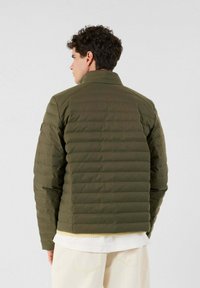 Hombre de espaldas con una chaqueta acolchada verde oliva sobre una camisa blanca y pantalones crema, frente a un fondo claro y liso.