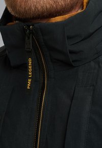 Zwarte jas met een hoge kraag, voorzien van een contrasterende oranje voering, ritssluiting en een gele logo label. Textuurstof en gedetailleerde stiksels.