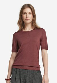 Ung kvinna med vågigt ljusbrunt hår som bär en kortärmad vinröd tröja och grå texturerade byxor mot en vit bakgrund.