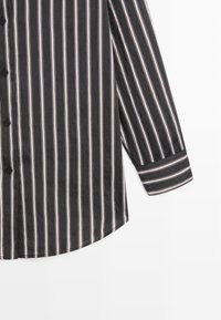 Czarna i biała koszula w paski z długimi rękawami, z guzikami, zaokrąglonym dołem i kontrastującymi czerwonymi liniami między paskami. Lekki materiał.