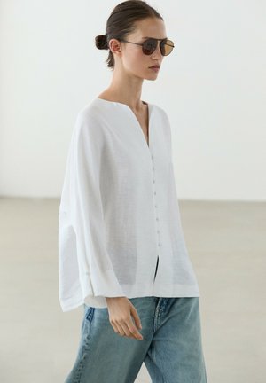 Frau mit dunkler Sonnenbrille, die eine lockere weiße Bluse mit Knöpfen und hellblaue Jeans trägt, steht vor einem einfarbigen hellen Hintergrund.