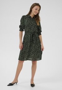 Robe chemise à motifs verts avec taille cintrée, manches courtes bouffantes et col. Portée avec des mules noires avec un détail de nœud sur le bout.