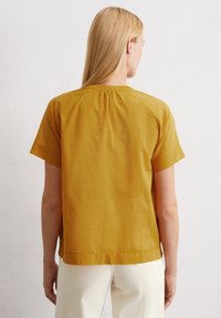 Senfgelbes Kurzarmoberteil mit lässigem Schnitt, das über ein gerafftes Rückendetail und einen glatten, leichten Stoff verfügt. Kombiniert mit weißen Hosen.
