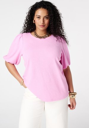 Vrouw met krullend haar, gekleed in een lichtroze top met geplooide mouwen, witte broek en gouden sieraden, poserend met één hand in de zak.