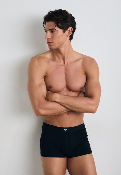 Boxers noirs ajustés avec un design moulant, en tissu doux, et une taille élastique arborant un petit logo à l'avant. Le modèle pose les bras croisés.