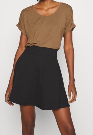 Vrouw draagt een bruine korte mouw t-shirt, ingestopt in een zwarte A-lijn rok met hoge taille, staand tegen een witte achtergrond.