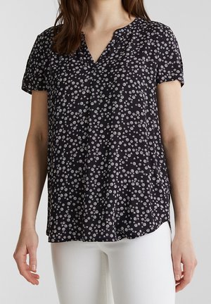 Femme portant un chemisier noir à manches courtes avec de petits motifs floraux blancs et un pantalon blanc, debout devant un fond uni.