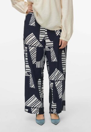 Pantaloni a gamba larga blu navy con motivi astratti bianchi. Il tessuto ha una finitura strutturata. Accoppiati con un top beige largo e scarpe con tacco blu.