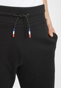 Zwarte katoenen joggers met ribgebreide tailleband, voorzien van een trekkoord met rode, witte en blauwe uiteinden. Diepe zijzakken inbegrepen.