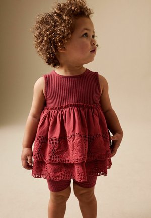 Toddler with curly hair wearing a sleeveless, layered maroon dress and matching shorts, looking to the right against a plain background.