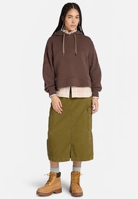 Bruine cropped hoodie met trekkoord, gedragen over een licht geruit overhemd. Olijfgroene knielange rok met cargozakken, gecombineerd met beige laarzen.
