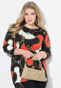 Langärmliges Oberteil in Schwarz, Rot und Weiß mit einem Kettenmuster; kombiniert mit einer goldenen, mit Pailletten besetzten Handtasche. Das Model hält die Handtasche.