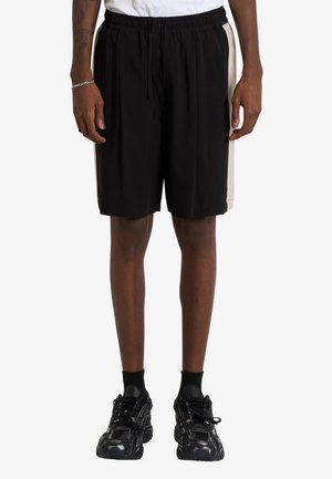 Persona che indossa pantaloncini sportivi neri con pannelli laterali bianchi, calzini neri e scarpe da corsa nere, in piedi contro uno sfondo semplice.