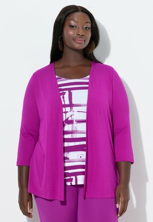 Kvinde med mørk hud og langt hår iført store store øreringe, en lilla cardigan med åben front, en mønstret hvid og lilla top samt lilla bukser.